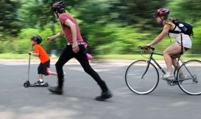 På billedet ses en far med sine to børn. Piger cykler, drengen løber på løbebånd og far løber med på sidelinjen. De har alle cykelhjelm på. 