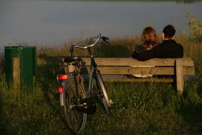 På billedet ses et ungt par, der sidder på en bænk i naturen. De har ryggen til kameraet. Tættest på kameraet står en damecykel parkeret