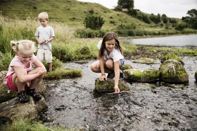 På billedet ses tre børn, der leger i vandkanten. En dreng står i baggrund og to piger sidder på hug i forgrunden- De står på to sten, der ligger i vandet