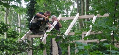 På billedet ses to mænd i færd med at bygge 'skelettet' til et hus i en skov