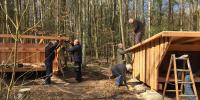 Billedet viser en flok mænd i færd med at bygge et træshelters i skoven
