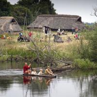 På billedet ses en masse gamle huse med stråtag i baggrunden i forgrunden sejler en mand en kano med tre børn på en sø