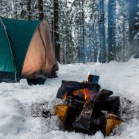 Billedet viser et telt, der er slået op i en snefyldt skov. foran teltet er der tændt et bål